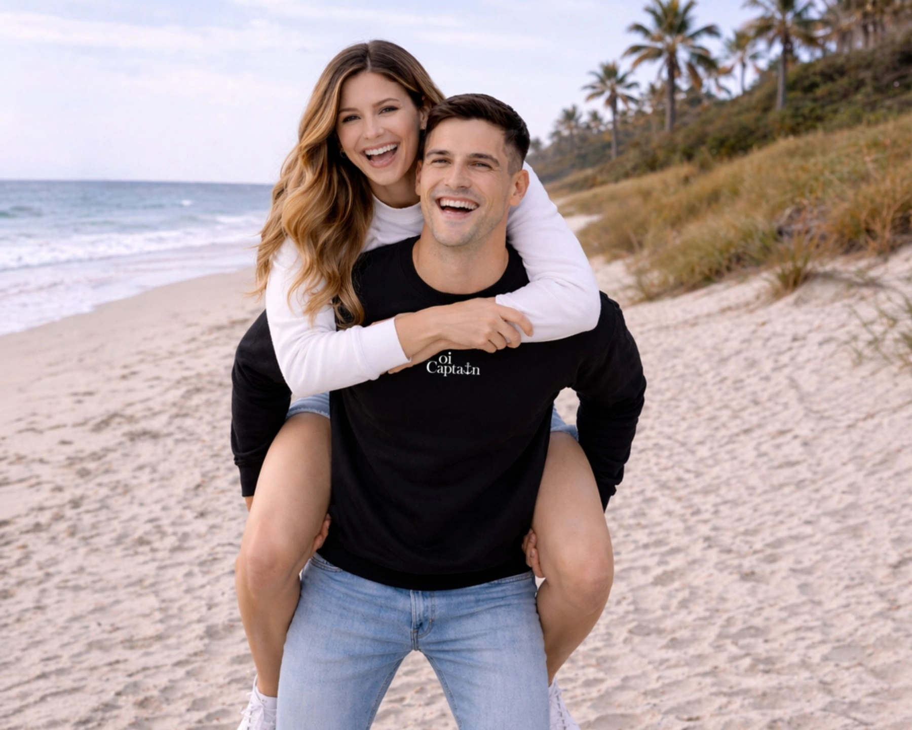 male model wearing black Oi Captain Dolce Vita Crew Neck Sweatshirt  piggybacking female model wearing white Oi Captain Dolce Vita Crew Neck Sweatshirt front view on a beach with palm trees in the background.jpg